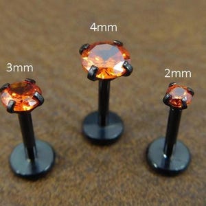 May include: Three black lip piercing studs with orange gemstones. The studs are 2mm, 3mm, and 4mm in size. The gemstones are held in place by four black prongs.