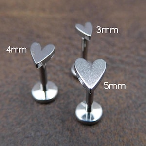 Könnte beinhalten: Drei silberfarbene Lippenpiercing-Stecker in Herzform. Die Stecker sind 3 mm, 4 mm und 5 mm groß. Der Schmuck wird auf einer braunen Oberfläche präsentiert.