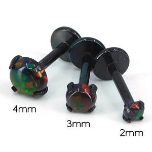 May include: Three black lip piercing studs with iridescent opal-like stones. The studs are 4mm, 3mm, and 2mm in size. The stones have a mix of green, red, and orange colors. The posts and flat backs are black.