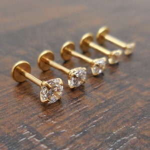 K&ouml;nnte beinhalten: F&uuml;nf goldfarbene Ohrstecker mit klaren, quadratisch geschliffenen Zirkonia-Steinen. Die Ohrringe sind in einer Reihe angeordnet, wobei die funkelnden Steine und das polierte Goldmetall im Mittelpunkt stehen. Der Hintergrund ist eine dunkle Holzoberfl&auml;che.