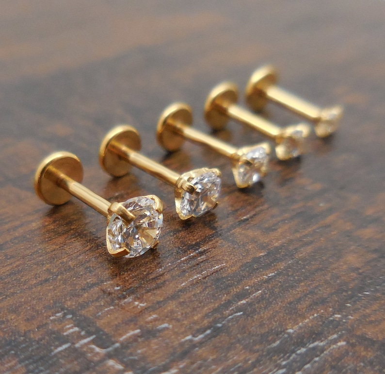 K&ouml;nnte beinhalten: F&uuml;nf goldfarbene Ohrstecker mit klaren, quadratisch geschliffenen Zirkonia-Steinen. Die Ohrringe sind in einer Reihe angeordnet, wobei die funkelnden Steine und das polierte Goldmetall im Mittelpunkt stehen. Der Hintergrund ist eine dunkle Holzoberfl&auml;che.