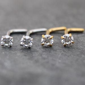 May include: Close-up of four nose studs with clear, round-cut gemstones. Two studs are silver-toned, and two are gold-toned. The gemstones are set in a four-prong setting, and the posts are L-shaped.