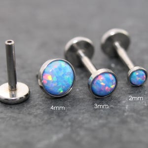 May include: A collection of silver-colored labret studs with blue opal-like stones. The studs are available in 2mm, 3mm, and 4mm sizes. The opal stones have a mix of blue, pink, and purple hues. The labret posts are straight and have a flat back.