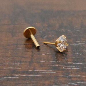 K&ouml;nnte beinhalten: Zwei goldfarbene Ohrstecker. Einer hat eine flache, runde R&uuml;ckseite mit einem Stift. Der andere Ohrring hat einen herzf&ouml;rmigen, klaren Edelstein, der in einer goldfarbenen L&uuml;nette gefasst ist. Die Ohrringe werden auf einer dunkelbraunen Holzoberfl&auml;che pr&auml;sentiert.