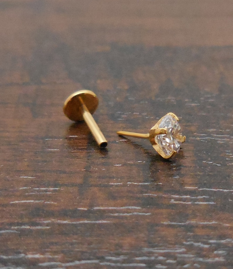 K&ouml;nnte beinhalten: Zwei goldfarbene Ohrstecker. Einer hat eine flache, runde R&uuml;ckseite mit einem Stift. Der andere Ohrring hat einen herzf&ouml;rmigen, klaren Edelstein, der in einer goldfarbenen L&uuml;nette gefasst ist. Die Ohrringe werden auf einer dunkelbraunen Holzoberfl&auml;che pr&auml;sentiert.
