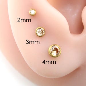 May include: Three gold-tone stud earrings with clear round stones, labeled with sizes 2mm, 3mm, and 4mm. Each earring features a bezel setting holding a single faceted stone, arranged vertically to demonstrate the different diameter options available.
