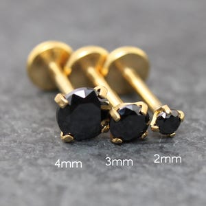 May include: Three gold-coloured labret studs with black gemstones. The studs are 4mm, 3mm, and 2mm in size. The gemstones are round and faceted, and the posts have a flat disc backing.