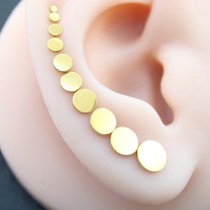 May include: A gold-toned ear climber earring featuring a series of nine circular discs that gradually increase in size from the top to the bottom, following the natural curve of the ear cartilage in a smooth, polished, and metallic finish.