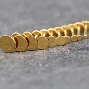 May include: A collection of gold-colored metal flat-back studs arranged in a row, each featuring a circular disc top with a textured, hammered finish and a straight post extending from the center of the back of each individual piece.
