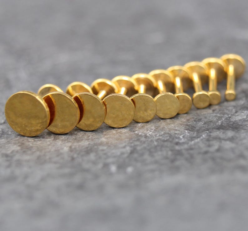 May include: A collection of gold-colored metal flat-back studs arranged in a row, each featuring a circular disc top with a textured, hammered finish and a straight post extending from the center of the back of each individual piece.