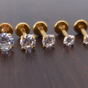 K&ouml;nnte beinhalten: F&uuml;nf goldfarbene Labret-Ohrringe mit klaren, rund geschliffenen Zirkonia-Steinen. Die Ohrringe sind in einer Reihe angeordnet, wobei die Steine in der Gr&ouml;&szlig;e abnehmen. Die Stifte und flachen R&uuml;ckseiten sind sichtbar.
