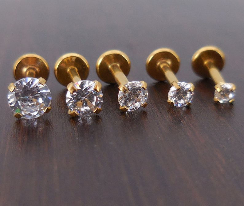 K&ouml;nnte beinhalten: F&uuml;nf goldfarbene Labret-Ohrringe mit klaren, rund geschliffenen Zirkonia-Steinen. Die Ohrringe sind in einer Reihe angeordnet, wobei die Steine in der Gr&ouml;&szlig;e abnehmen. Die Stifte und flachen R&uuml;ckseiten sind sichtbar.