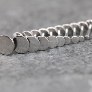 May include: A collection of silver-toned metal labret studs arranged in a row, each featuring a flat, circular disc top and a straight post. The studs vary in size, with the largest disc positioned in the front and the smallest at the back.