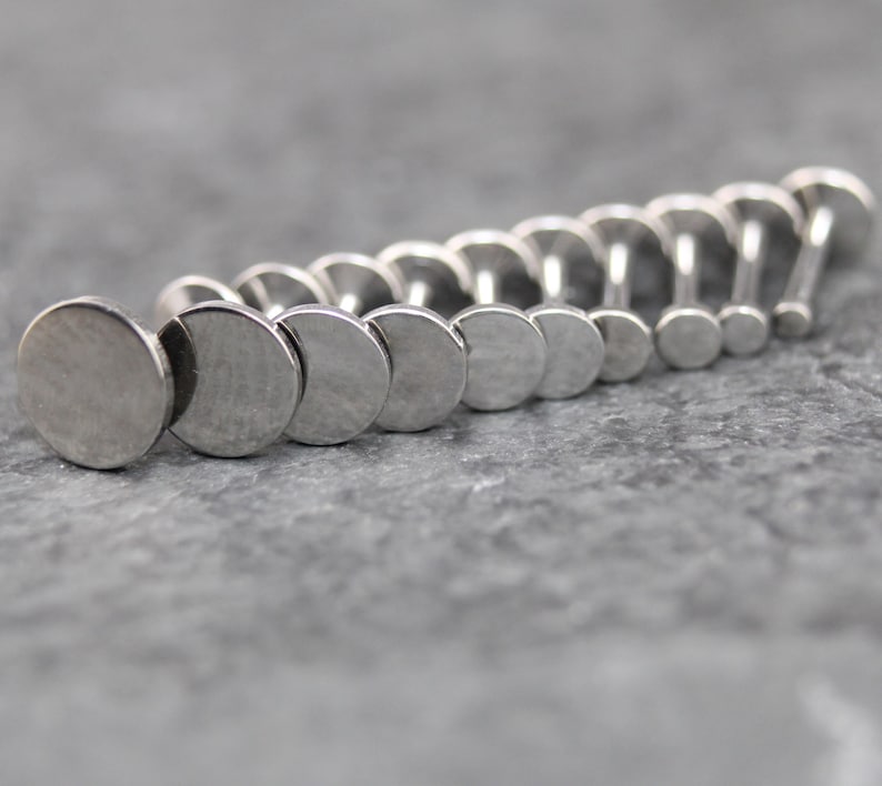 May include: A collection of silver-toned metal labret studs arranged in a row, each featuring a flat, circular disc top and a straight post. The studs vary in size, with the largest disc positioned in the front and the smallest at the back.
