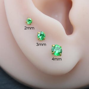 May include: Three round, prong-set green gemstone stud earrings in gold-tone settings, displayed on an ear model. The studs are labeled with their respective sizes: 2mm, 3mm, and 4mm, showing the scale of each individual piece clearly.