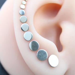 May include: A series of nine silver-toned circular stud earrings arranged in a graduated size sequence along the outer edge of an ear. Each earring features a smooth, flat, polished metallic surface, starting from a tiny dot at the top to a larger disc at the base.