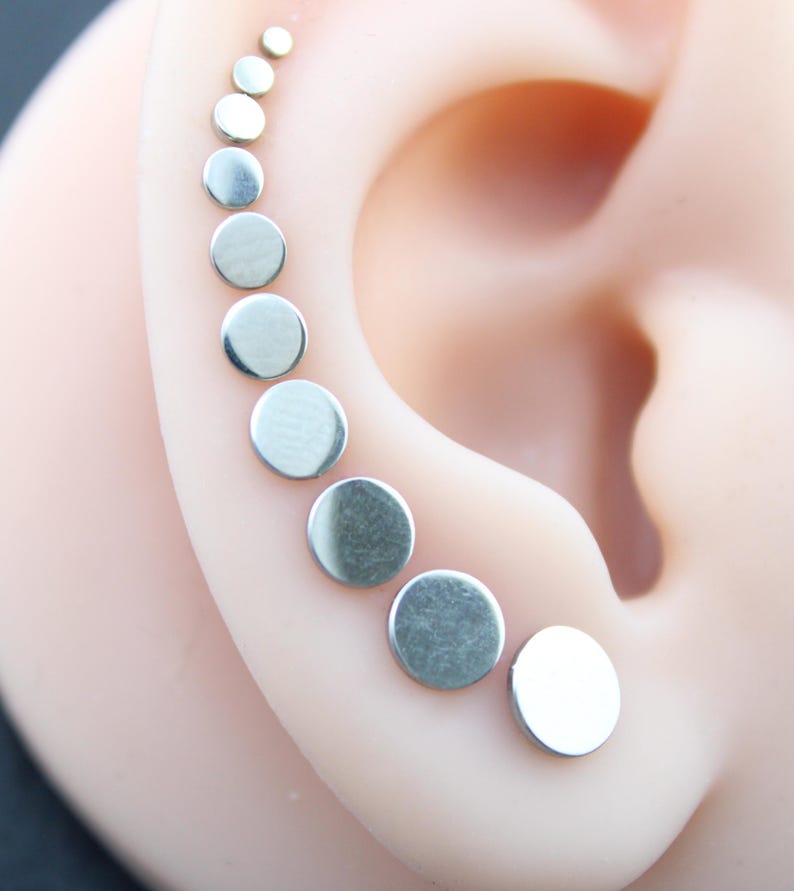 May include: A series of nine silver-toned circular stud earrings arranged in a graduated size sequence along the outer edge of an ear. Each earring features a smooth, flat, polished metallic surface, starting from a tiny dot at the top to a larger disc at the base.