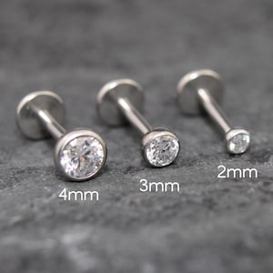 May include: Three silver-toned labret studs with clear, round-cut gemstones in bezel settings. The studs are labeled with sizes 4mm, 3mm, and 2mm, indicating the diameter of the gemstone. Each piece features a flat circular back disc.