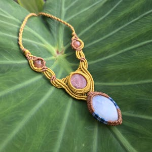 May include: A macrame necklace featuring a mustard yellow cord woven into intricate patterns. The design includes two small round reddish-brown stones, one central oval reddish-brown stone, and a large teardrop-shaped light blue stone at the base.