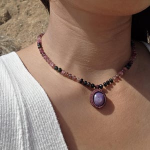 May include: A necklace featuring a central oval purple cabochon stone encased in a dark purple woven frame, suspended from a strand of small, faceted beads in alternating shades of translucent pink, deep red, and opaque black.