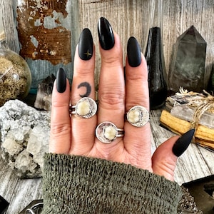 Puede incluir: Tres anillos de plata con incrustaciones de piedras blancas. Los anillos se llevan en los dedos de una mano con esmalte de uñas negro mate.