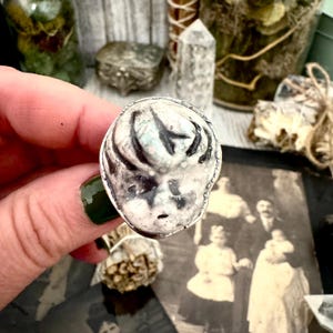 May include: A silver ring with a detailed doll's face design. The face is predominantly white with black detailing, creating a stark contrast. The ring is held in a hand with dark green nail polish. A vintage photograph is visible in the background.