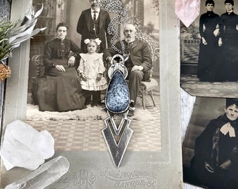 May include: A silver pendant necklace with a gray and white teardrop-shaped stone. The pendant is in the shape of a stylized arrow with a V-shaped design. The necklace is hanging in front of a vintage photograph of a family.