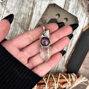 May include: A silver necklace with a clear quartz crystal pendant and a purple gemstone accent. The pendant is wrapped in silver wire and has a textured finish.