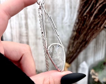 May include: A silver-toned pendant necklace featuring a teardrop-shaped, clear stone with gray inclusions. The pendant has a geometric design with a chain and is held up by a finger with a black nail.