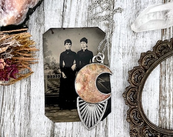 May include: A silver chain necklace with a large, crescent moon pendant. The pendant is made of metal with a textured, pink and gold finish. The pendant is hanging over an antique photograph of two women.