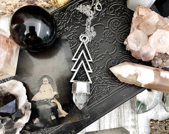 May include: A silver necklace with a clear quartz crystal pendant shaped like a triangle. The pendant is layered with three triangles, creating a geometric design. The necklace is displayed on a black surface with other crystals and a vintage photograph.