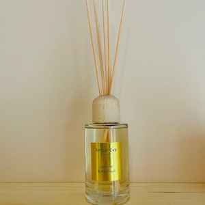 Puede incluir: Un difusor de aromaterapia de vidrio transparente con varillas de madera. La botella tiene una tapa de madera y una etiqueta dorada que dice "Amber Eve London" y "Jasmine & Patchouli". El difusor está sobre una estantería de madera clara.