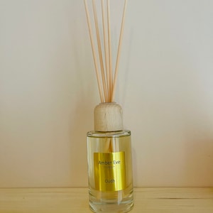 Op de afbeelding: Een heldere glazen diffuserfles met een houten dop en rietjes. De fles heeft een gouden label met de tekst "Amber Eve London" en "Oudh". De rietjes zijn lichtbruin en steken omhoog.