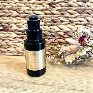 Puede incluir: Una botella de vidrio negro de Amber Eve Face Elixir Miracle Oil con un dispensador de bomba. La etiqueta es dorada con texto negro. Flores secas y un fondo tejido están en el fondo.