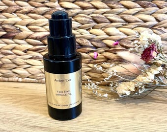 May include: A black glass bottle of Amber Eve Face Elixir Miracle Oil with a pump dispenser. The label is gold with black text. Dried flowers and a woven background are in the background.