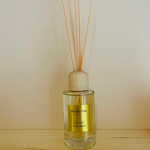 May include: A clear glass diffuser bottle with a wooden cap and reeds. The bottle has a gold label with the text "Amber Eve London" and "Myrrh & Tonka Bean". The reeds are light brown and stand upright.
