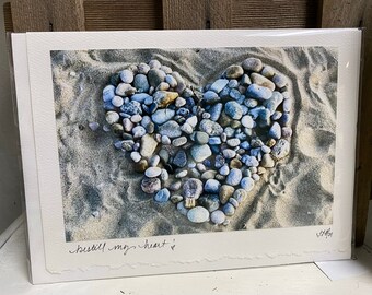 May include: A print featuring a heart shape made of smooth, colorful stones on a sandy beach. The heart is composed of various shades of blue, gray, and brown stones. The words "bestill my heart" are written in cursive at the bottom.