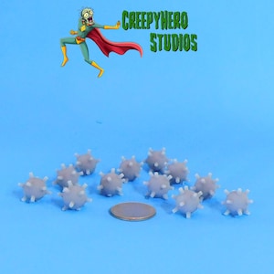 May include: A collection of small, gray, spiky objects, possibly miniature sculptures or game pieces, are arranged on a blue surface. A coin is placed near the objects for size comparison. The image also features a cartoon superhero and the text "CREEPYHERO STUDIOS".