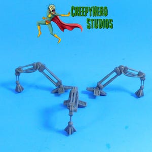May include: Three gray, mechanical-looking objects with articulated arms and decorative tassels. The objects are arranged on a blue background. The text "CREEPYHERO STUDIOS" is in green above the objects, along with a cartoon superhero.