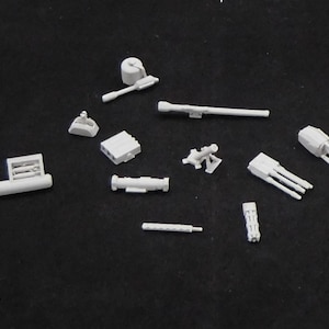 May include: A collection of small, light gray model military components. The assortment includes various miniature cannons, rocket launchers, and other detailed parts, likely for a scale model project. The pieces are arranged on a dark surface.