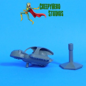 May include: A gray plastic toy spaceship and a stand are displayed against a blue background. The spaceship has a unique design with wings and a cockpit. The stand is hexagonal. The image also features a cartoon superhero and the text "CREEPYHERO STUDIOS."