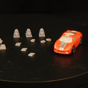 May include: A red toy race car with white stripes and a white design on the hood. The car is surrounded by small gray toy parts.