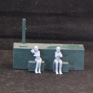 May include: Two gray plastic figurines of people sitting on a green platform. The figures are wearing helmets and are posed with their hands in their laps.