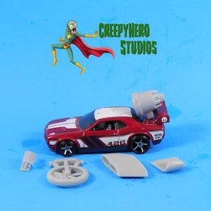 May include: A red toy car with a white roof and black racing stripes. The car has a gray spoiler and a gray engine on the back. The car is on a blue surface with a gray wheel and a gray piece of plastic.