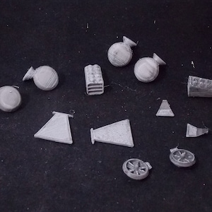 May include: A collection of small, white, 3D printed objects, including cones, wheels, and blocks. The objects are arranged on a dark surface.