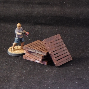 May include: A stack of four wooden pallets, painted brown, with a miniature figure standing nearby. The pallets are stacked in a staggered pattern, with the top pallet slightly offset from the others.