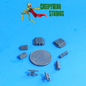 May include: A collection of small, gray plastic model parts, including rectangular and cylindrical shapes, and two larger components with detailed designs. A US quarter is included for size reference. The image also features a cartoon superhero and the text "CREEPYHERO STUDIOS".