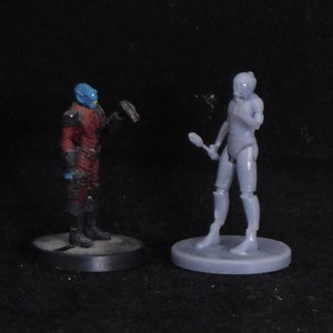 May include: Two painted miniature figures, one blue and red, the other gray, standing on circular bases. The blue and red figure is holding a gun, the gray figure is holding a staff.