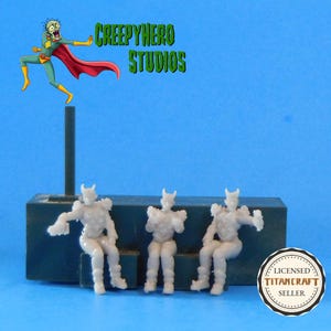 May include: Three small, light gray figurines of seated characters with horned helmets and shoulder armor. They are on a dark gray platform. The image includes the text "CREEPYHERO STUDIOS" and a cartoon character. A circular sticker reads "LICENSED TITANCRAFT SELLER."
