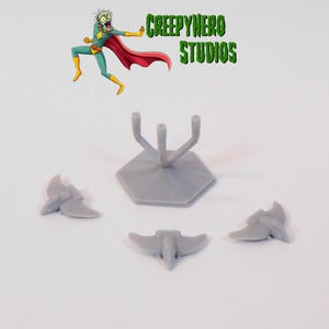 May include: A set of four gray plastic spaceship miniatures, including a stand with three prongs. The stand is hexagonal shaped. The miniatures are all the same design, with a pointed nose and two wings. The miniatures are on a white surface. The text "Creepyhero Studios" is visible in the background.